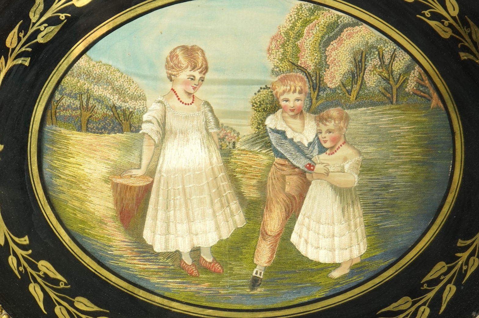 A large oval early 20th century embroidery of three children in a landscape. Worked in mostly stem stitch with knotting embroidery on the trees. The embroidery framed in Verre Eglomise glass and a wide wooden gilt frame